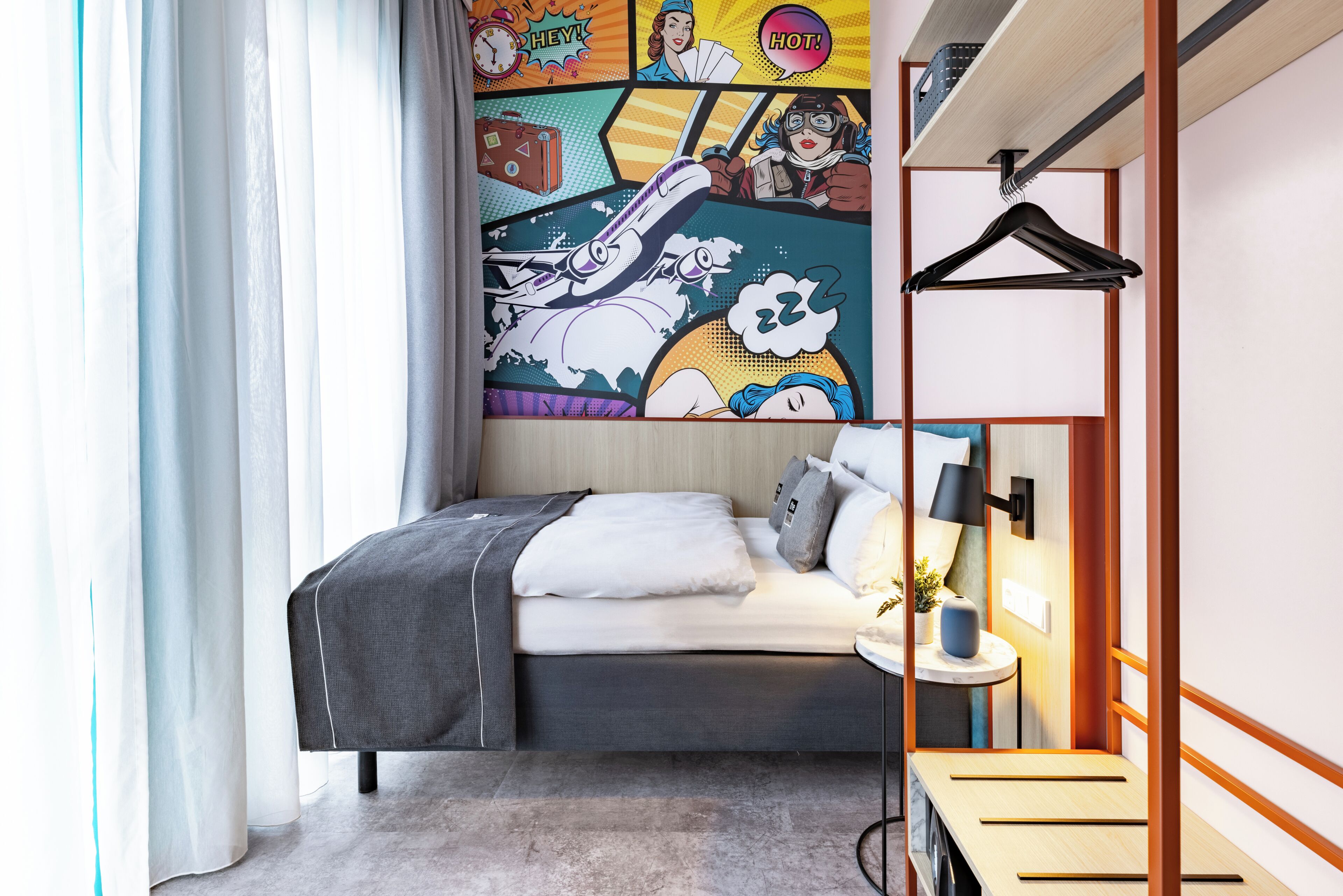 A modern hotel room with a bed, a bedside table with a lamp, a wall-mounted shelf, and a large comic-style mural.