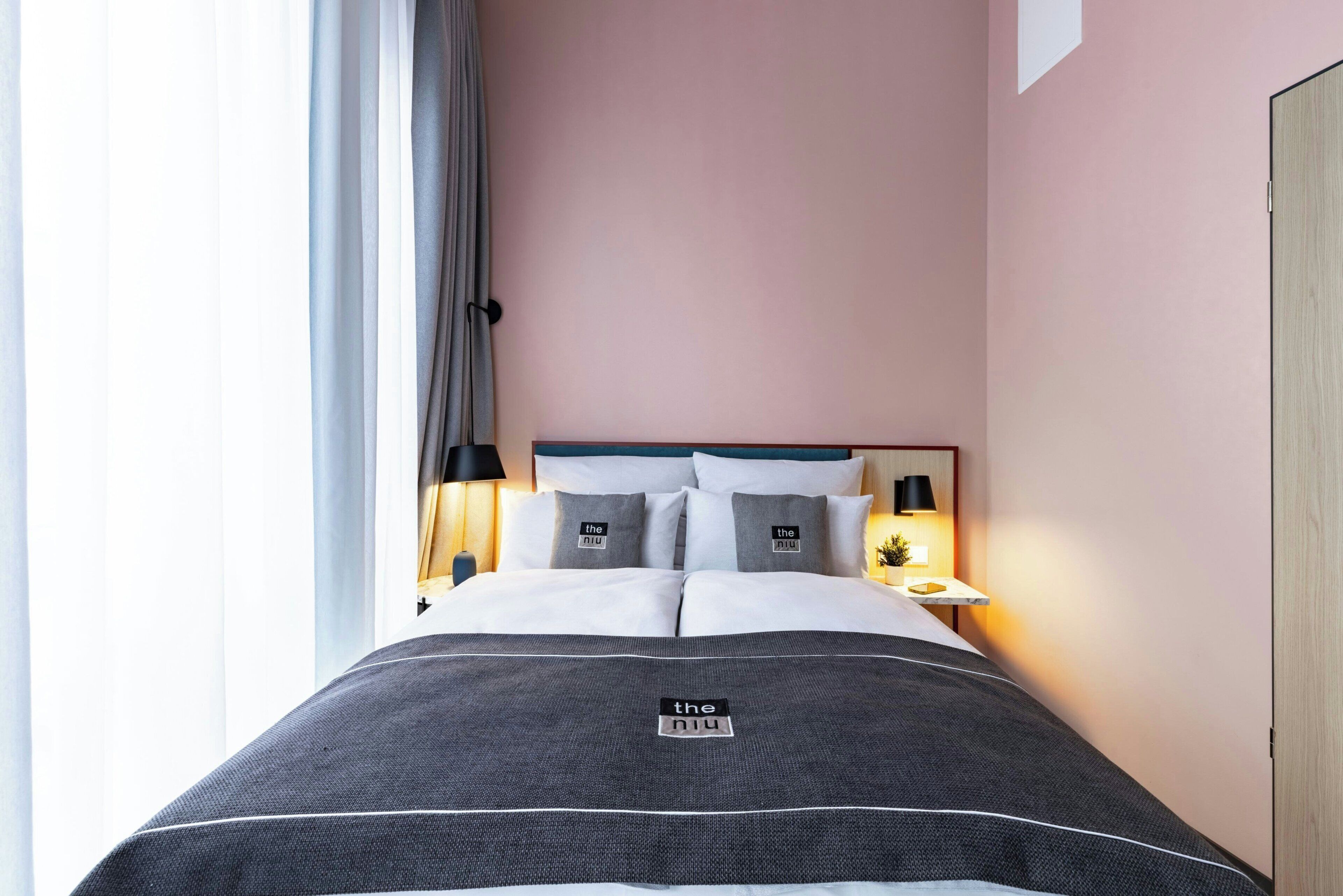 Habitació d'hotel amb un llit, una tauleta de nit, una làmpada i una finestra amb cortines.