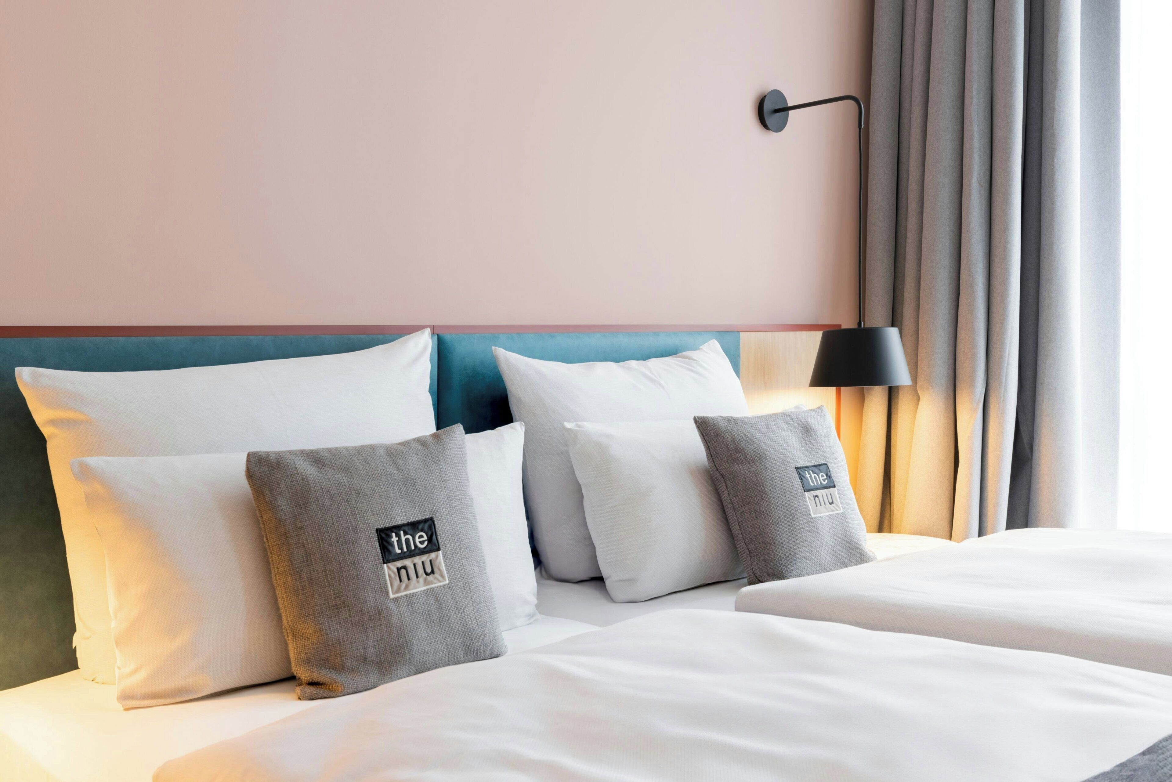 Ein Hotelzimmer mit Bett, Kissen, Kopfteil, einer Wandlampe und Vorhängen.