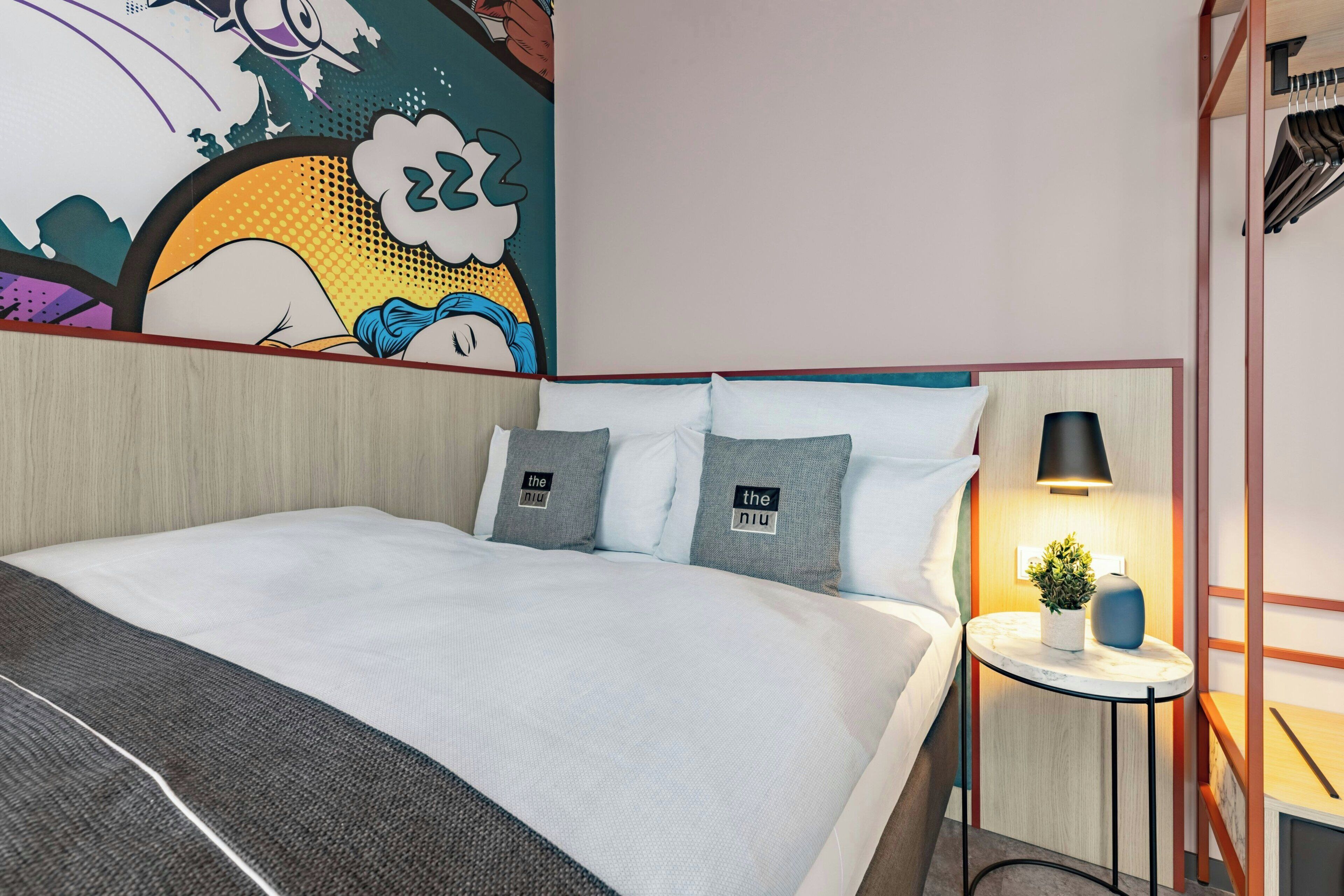 Moderni hotellihuone, jossa on suuri sänky, yöpöytä lampun kanssa ja värikäs seinämaalaus.