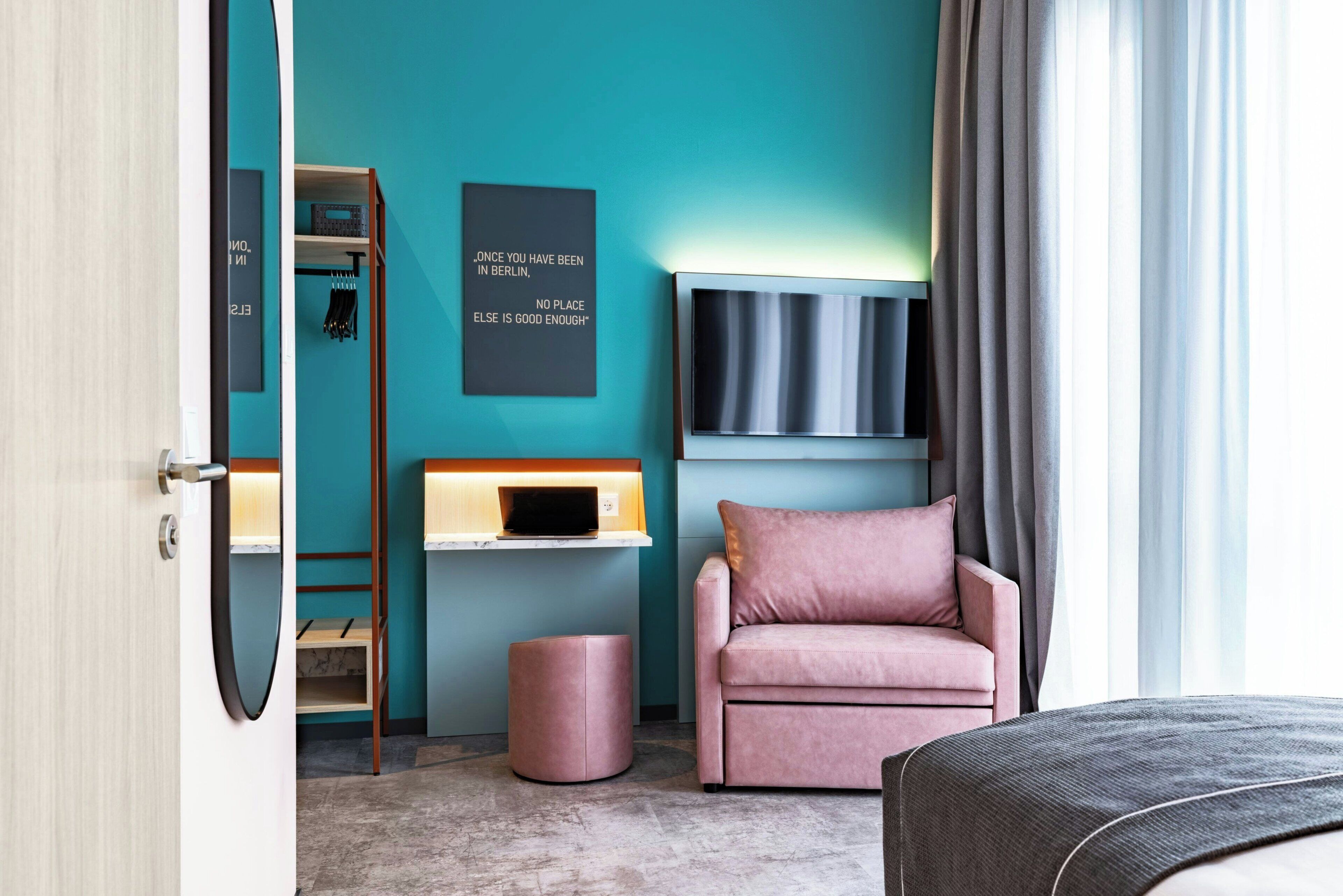 A modern hotel room with a teal accent wall, a flat-screen TV, a pink armchair, and a small wooden shelf.