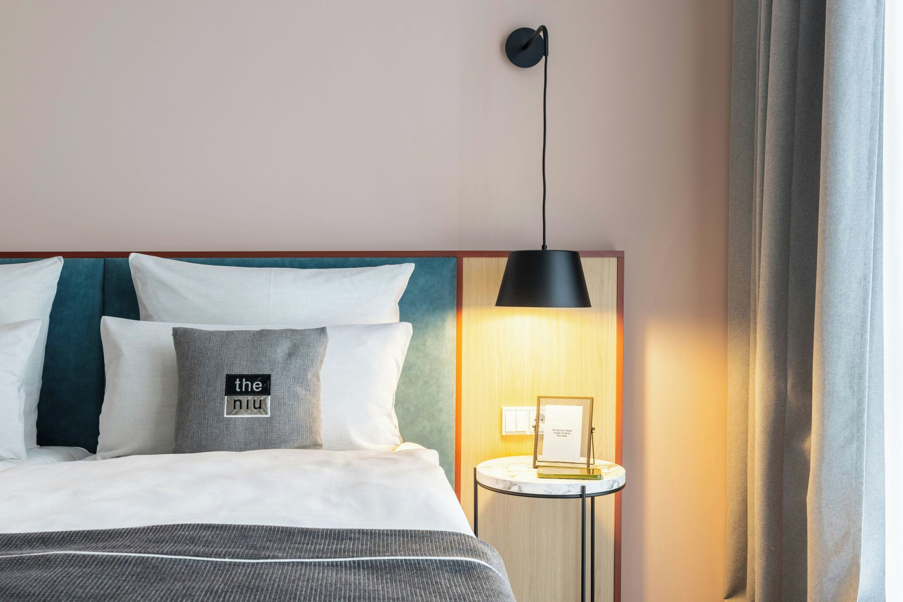 A hotel room with a bed, pillows, a bedside table with a lamp, and a wall-mounted light fixture.