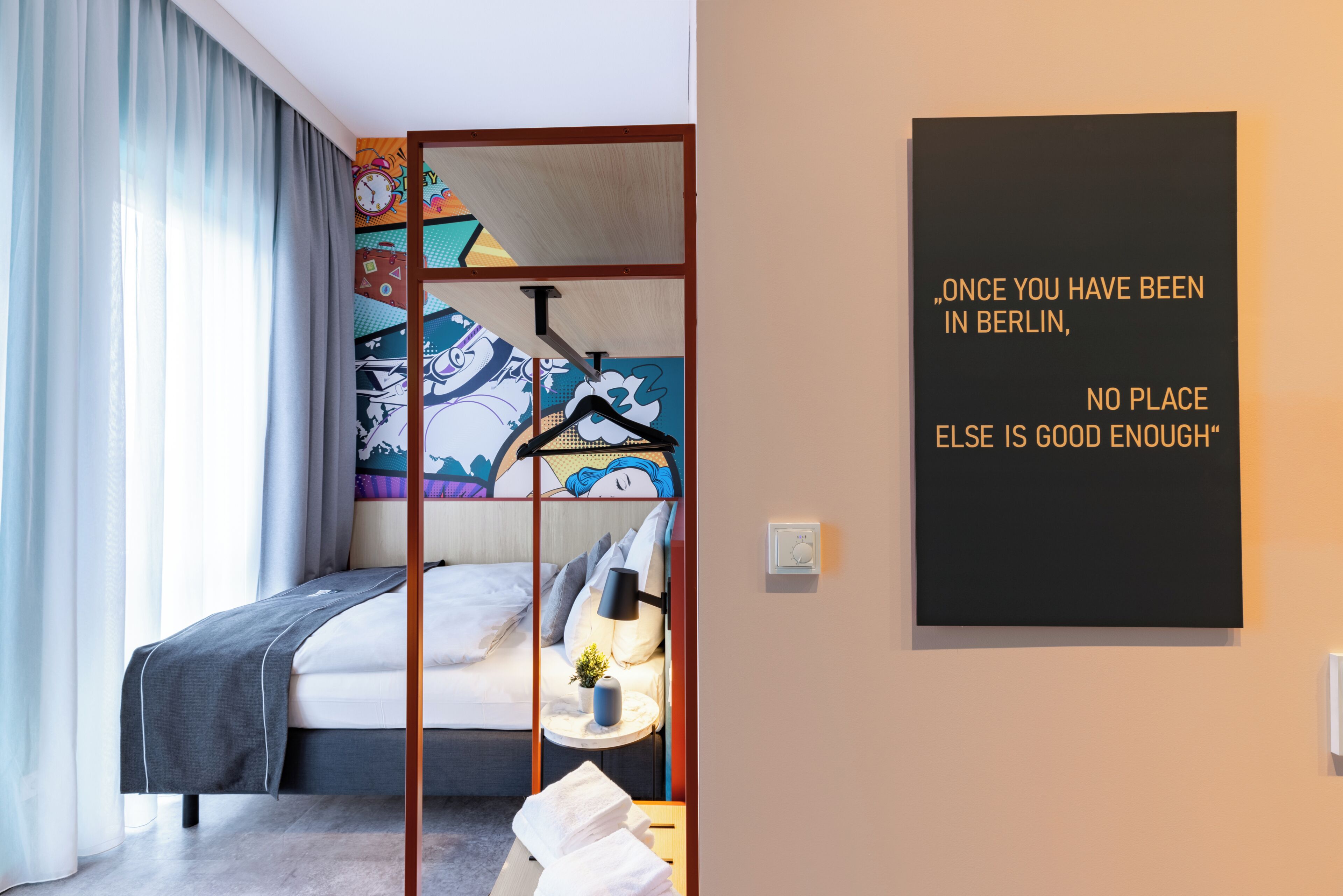 A hotel room with a bed, a side table, a wall art piece, and a window with curtains.