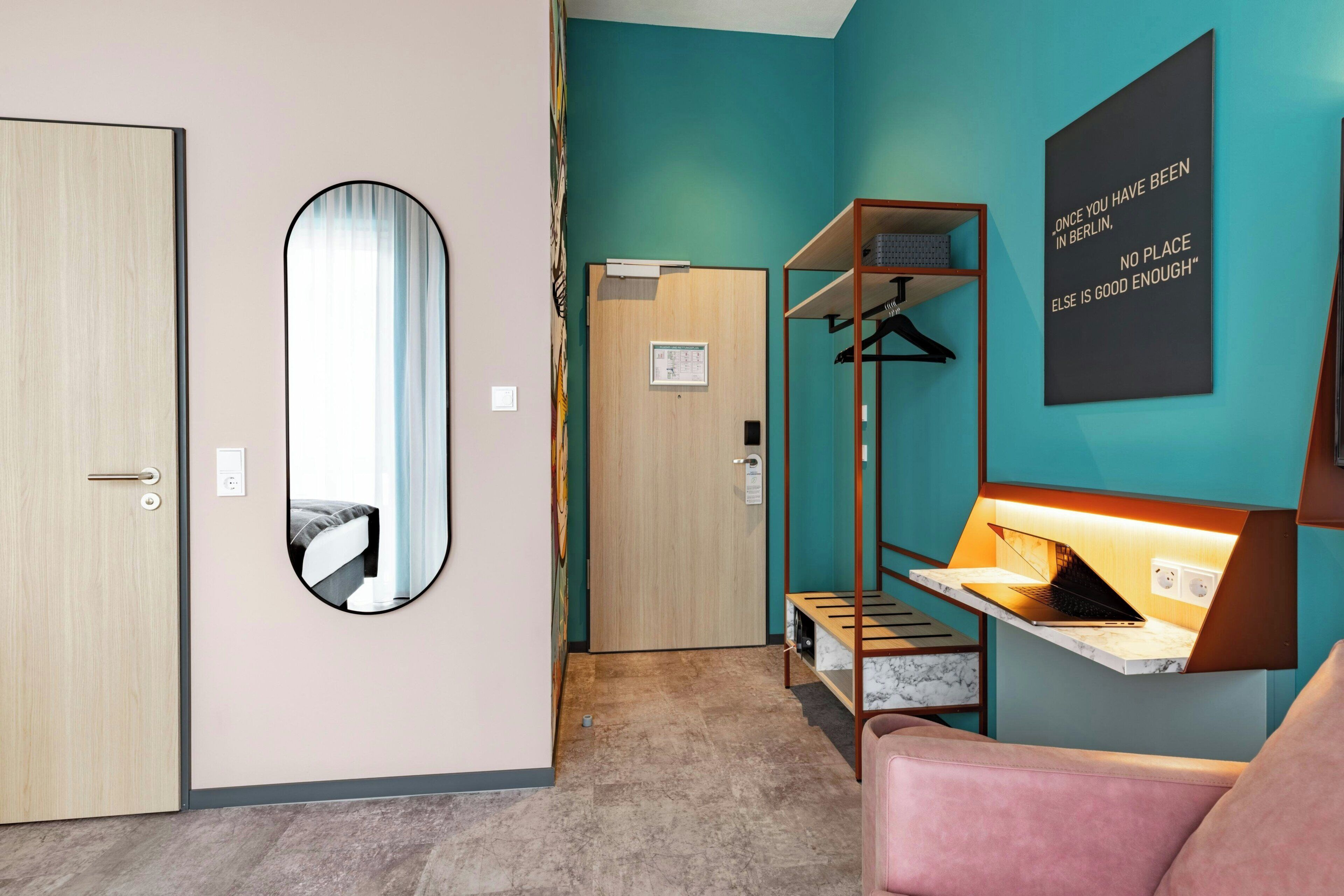 Une chambre d’hôtel avec une porte en bois, un miroir, un lit, un bureau avec un ordinateur portable, une étagère suspendue et une citation inspirante sur le mur.