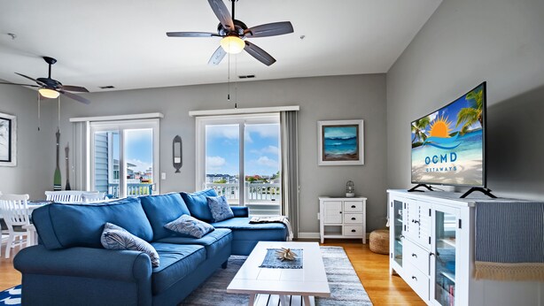 A modern living room with a blue sectional sofa, a white coffee table, a ceiling fan, a TV on a white cabinet, and a large window with a view of the city.