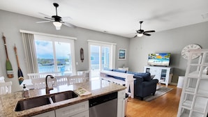 A modern kitchen with granite countertops, white cabinetry, and a dishwasher. There is a dining area with white chairs and a table, and a living room with a blue sofa and a television.