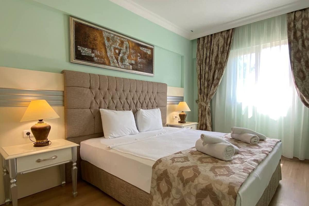 A hotel room with a large bed, two bedside tables with lamps, a headboard, a painting on the wall, and a window with curtains.