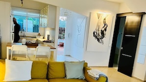 Una sala de estar moderna con un sofá amarillo, una mesa de centro de madera y una cocina visible a través de una puerta abierta.