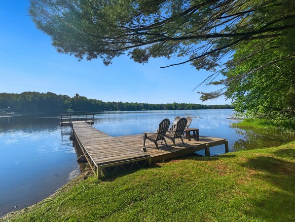 A wooden dock extending into calm water, with two Adirondack chairs and a small table on the dock.