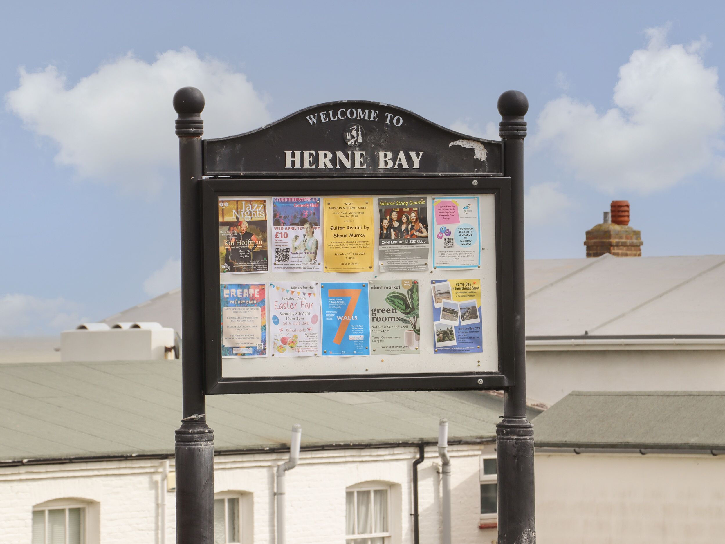 Señal de bienvenida a Herne Bay con un tablón de anuncios mostrando varios carteles de eventos.