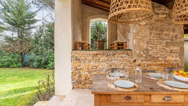 Un espacio de comedor al aire libre con un muro de piedra de fondo, una mesa de madera y luminarias tejidas.