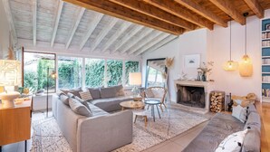 Amplia sala de estar con un sofá grande, una mesa de centro y una chimenea. Hay ventanas amplias que dejan entrar luz natural y ofrecen vistas al exterior.