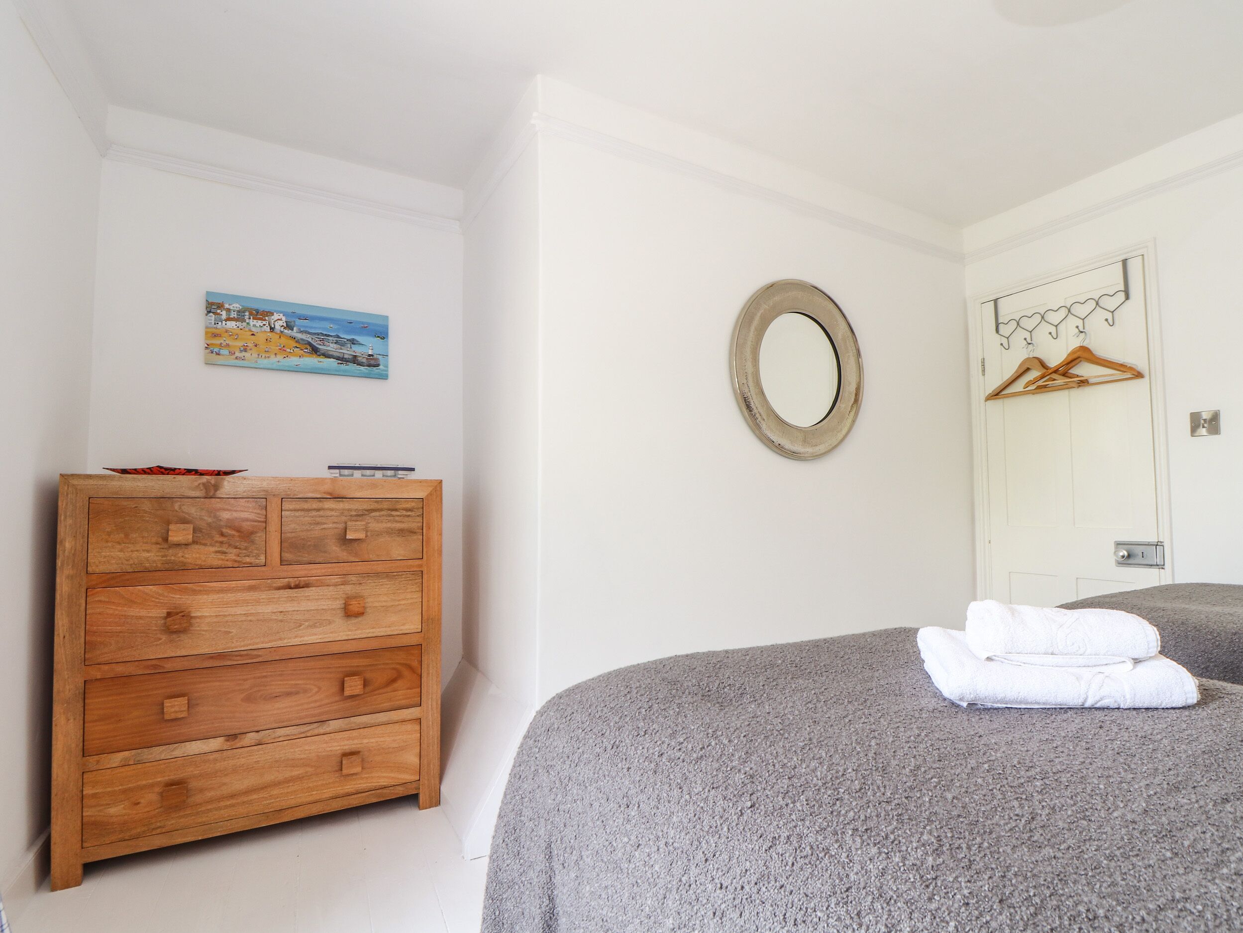 A bedroom with a wooden dresser, a round mirror, a bed with a grey blanket, and a door with a coat rack.