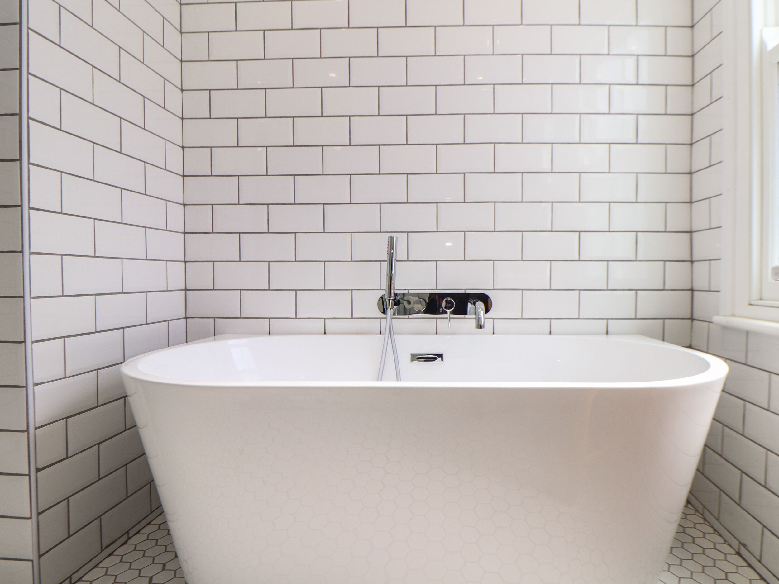 A modern bathroom with white subway tiles on the walls and floor, a freestanding bathtub, and a window on the right side.