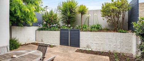 A patio area with a wooden table and chairs, surrounded by a low concrete wall with plants and a dark gate.