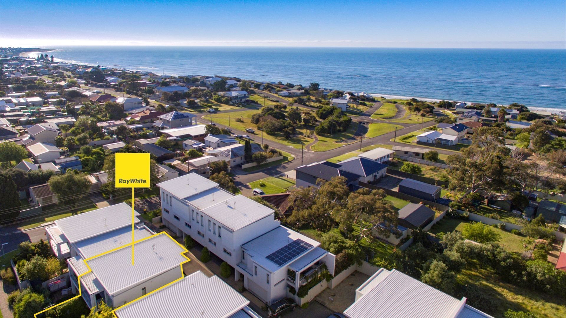 Vista aérea de uma área residencial com casas, um imóvel identificado como “RayWhite” e uma linha costeira ao fundo.