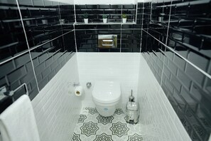 A bathroom with black and white patterned tiles, a white toilet, a towel rack, and a small shelf with plants. - Yörükali Tesisleri (Adalar)