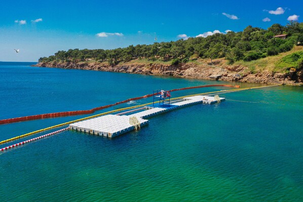 A pier extending into a clear blue sea with a rocky shoreline and trees in the background. - Yörükali Tesisleri (Adalar)