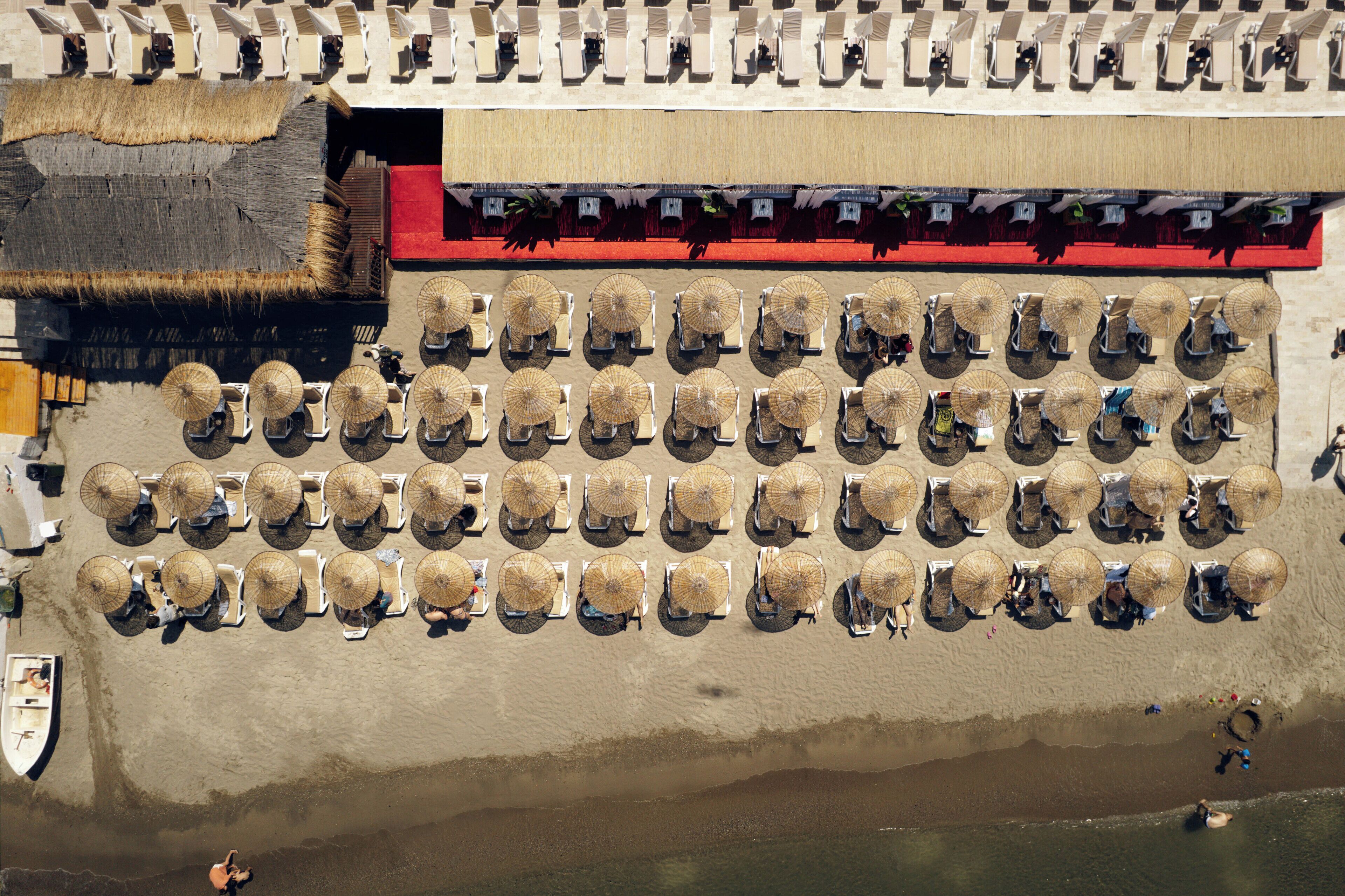 Vista aérea de una playa con hileras de sombrillas y sillas de playa, una estructura de techo de paja y un edificio con toldo rojo.