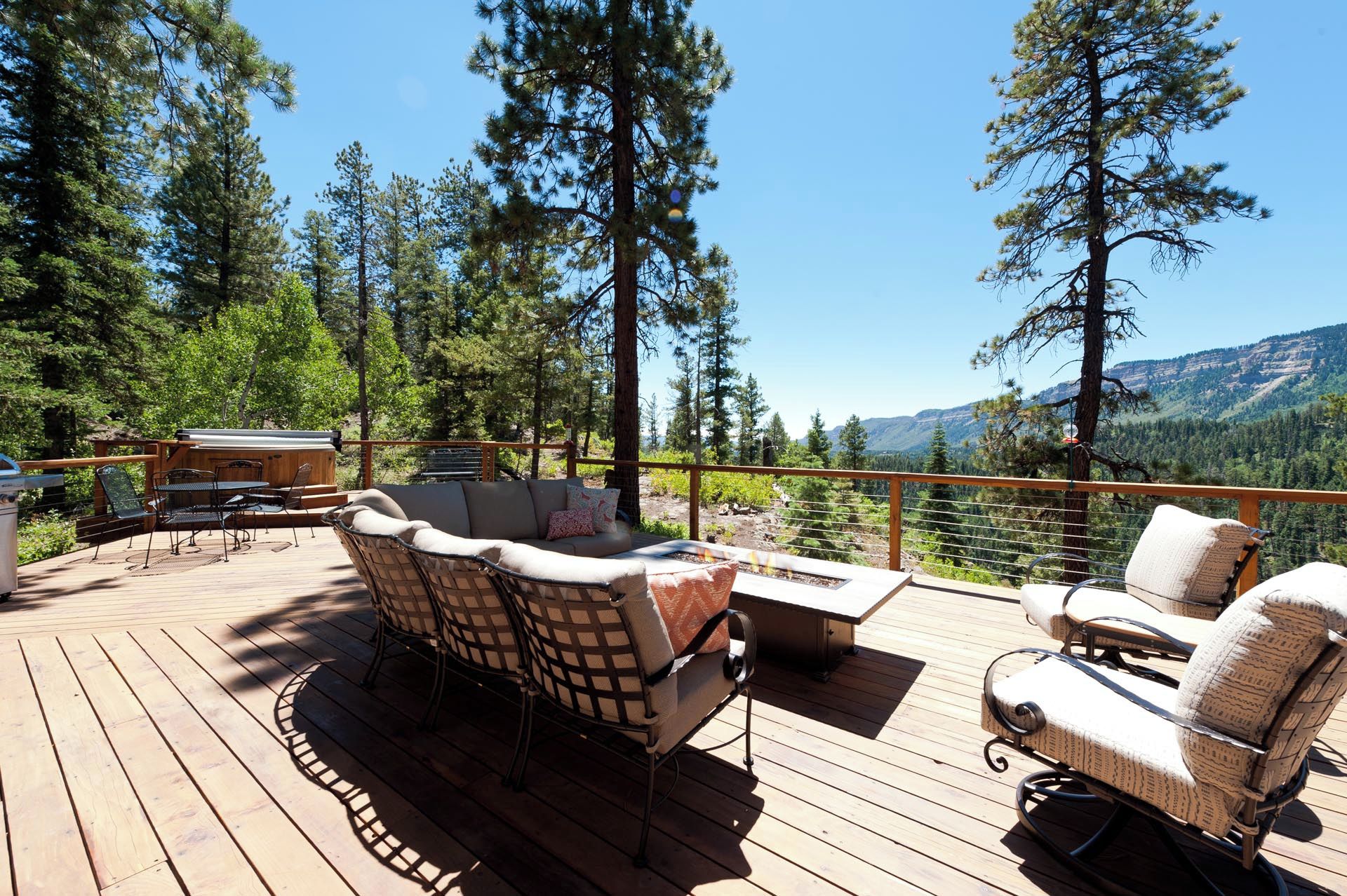 Une terrasse en bois, aménagée avec du mobilier d’extérieur, notamment un canapé et des fauteuils, qui offre une vue sur une zone boisée et, au loin, sur des montagnes.