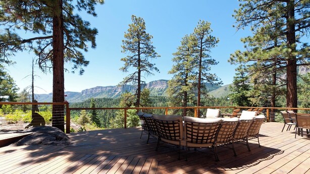 A wooden deck with outdoor furniture, surrounded by tall pine trees and a mountainous backdrop. - Luxury Home - Huge Deck - Great Views - Hot Tub/Fire Table - 2 Miles to Purg (Durango)