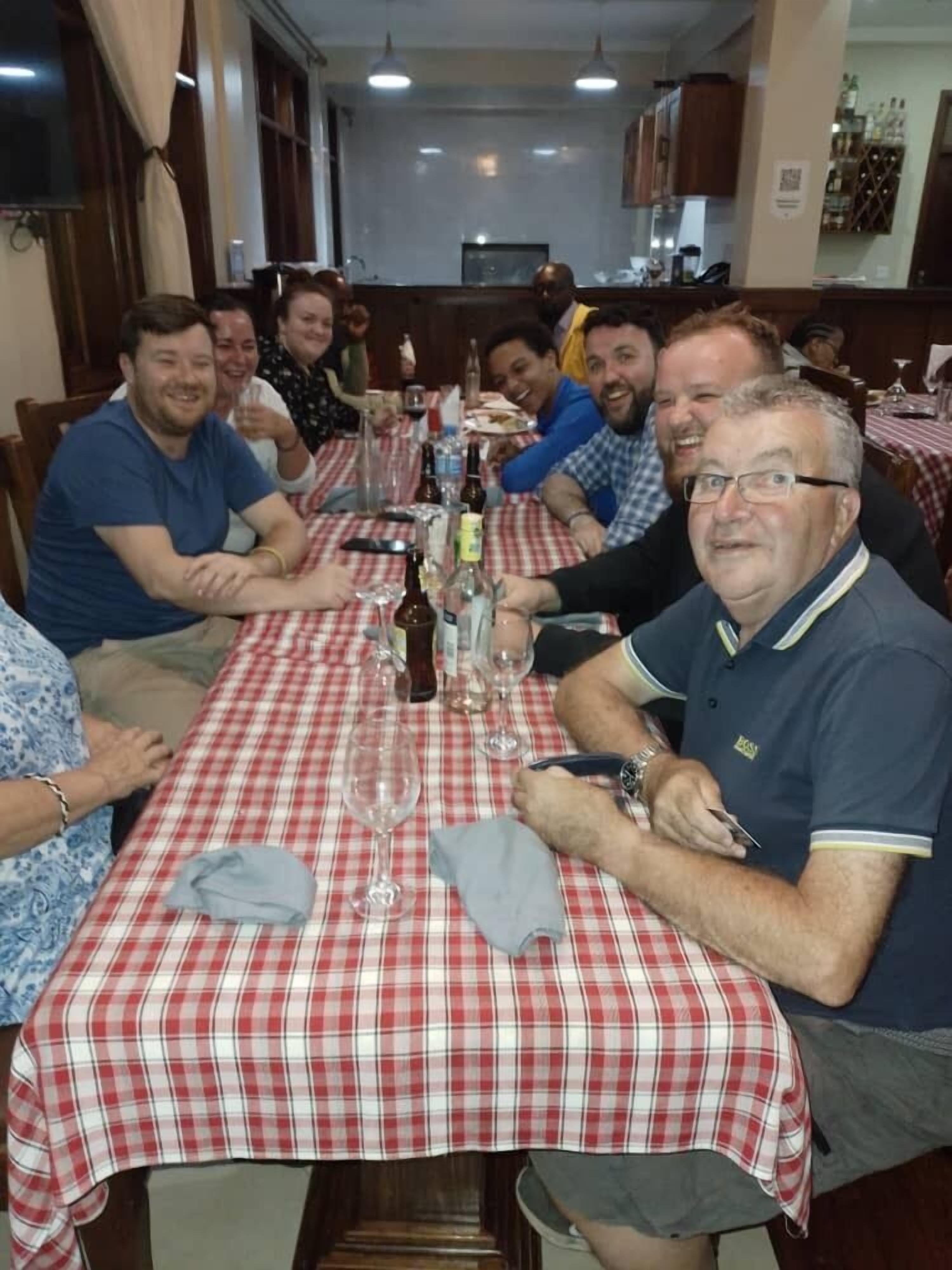 A group of people seated at a table with a red and white checkered tablecloth, in a restaurant setting with wooden furniture and a bar in the background.