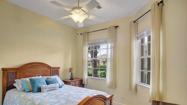 Ein Schlafzimmer mit einem Bett, einem Deckenventilator, einem Fenster mit Vorhängen und einem Nachttisch mit Lampe.