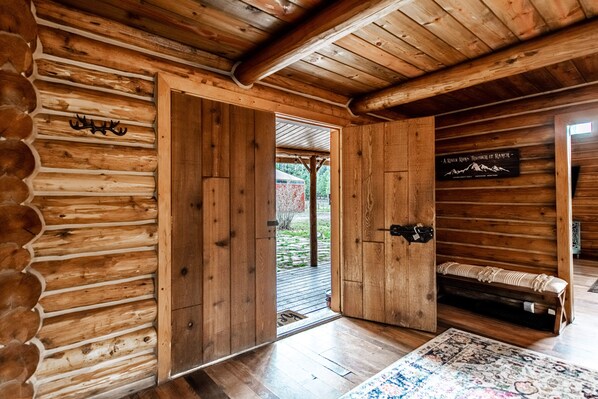 Interior de uma cabana de toras com uma porta de madeira que dá para o exterior, um banco com um cobertor e uma placa na parede.