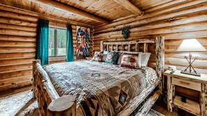 Ein Schlafzimmer in einer Blockhütte mit einem großen Bett, Holzwänden, einem Fenster mit Vorhängen und einem Nachttisch mit Lampe.