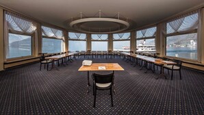 Een vergaderzaal met een grote ovale tafel, meerdere stoelen en grote ramen met uitzicht op een waterpartij en gebouwen.