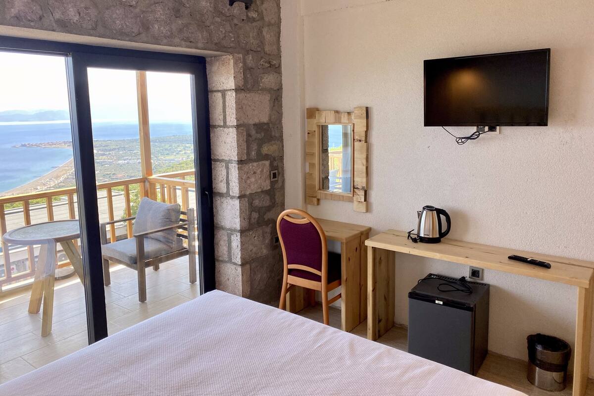 A hotel room with a balcony, a desk, a chair, a bed, a TV, and a small refrigerator.