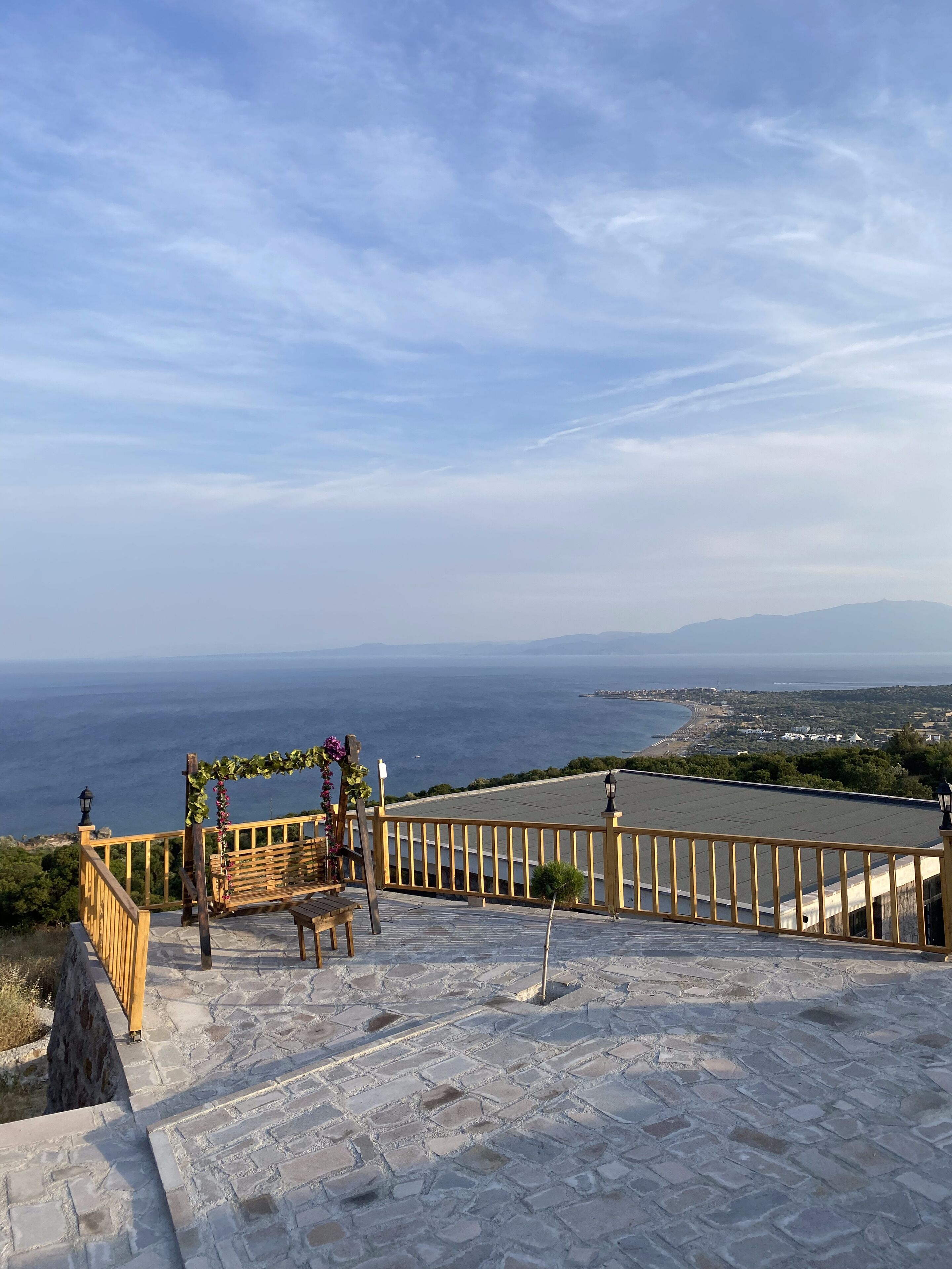 A stone-paved terrace with a wooden bench and a pergola adorned with flowers, overlooking a coastal landscape.