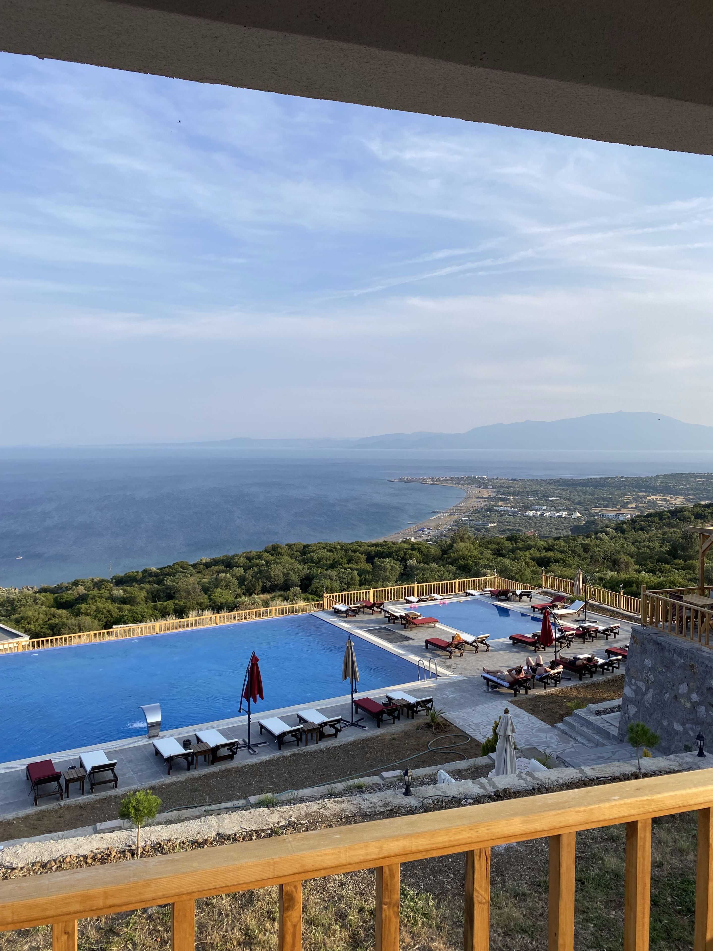A pool area with lounge chairs and umbrellas, surrounded by a wooden deck and a view of the sea and distant mountains.