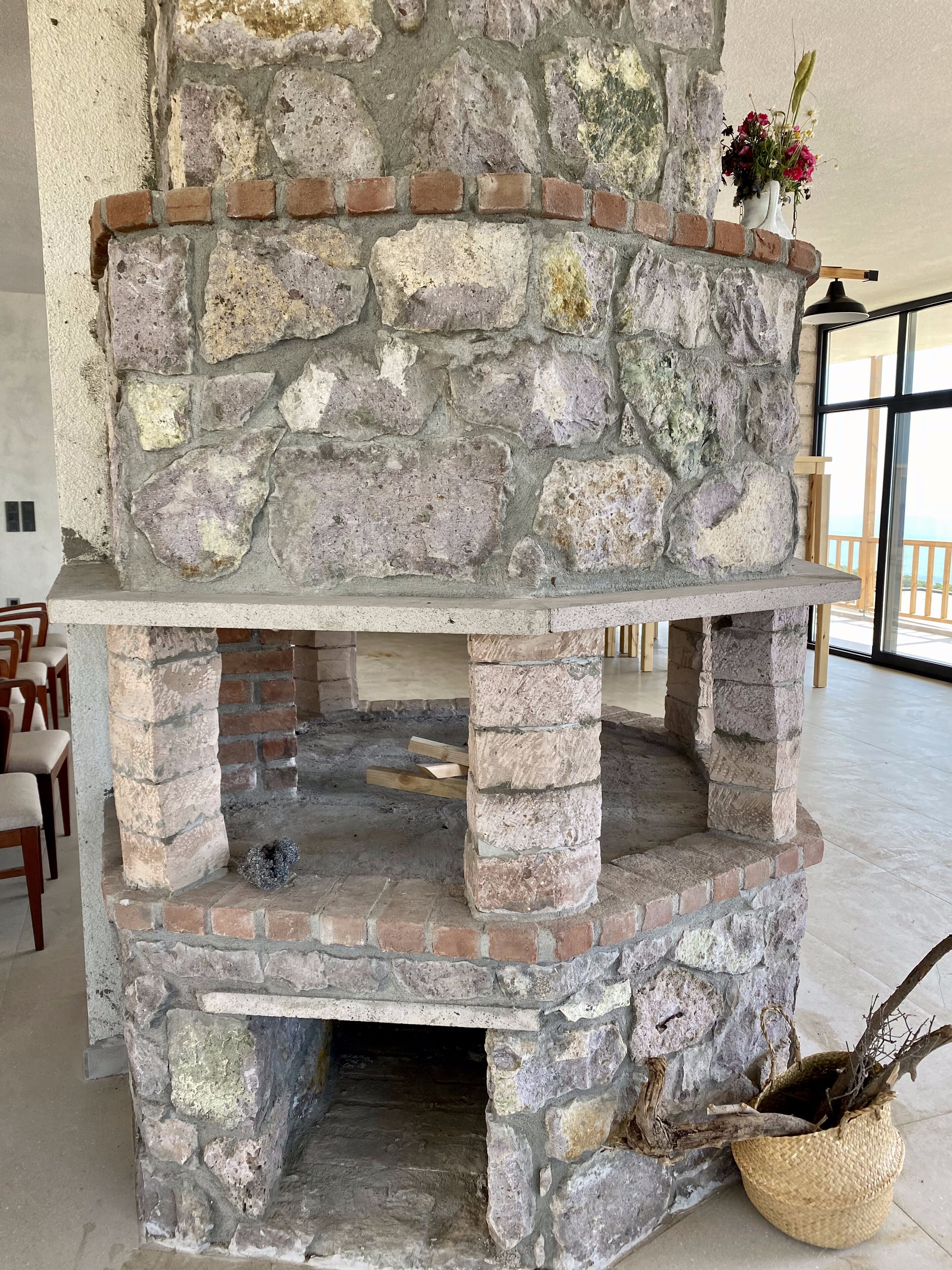 Una chimenea de piedra con marco de ladrillo, un florero encima y una cesta de mimbre con ramas en la base.