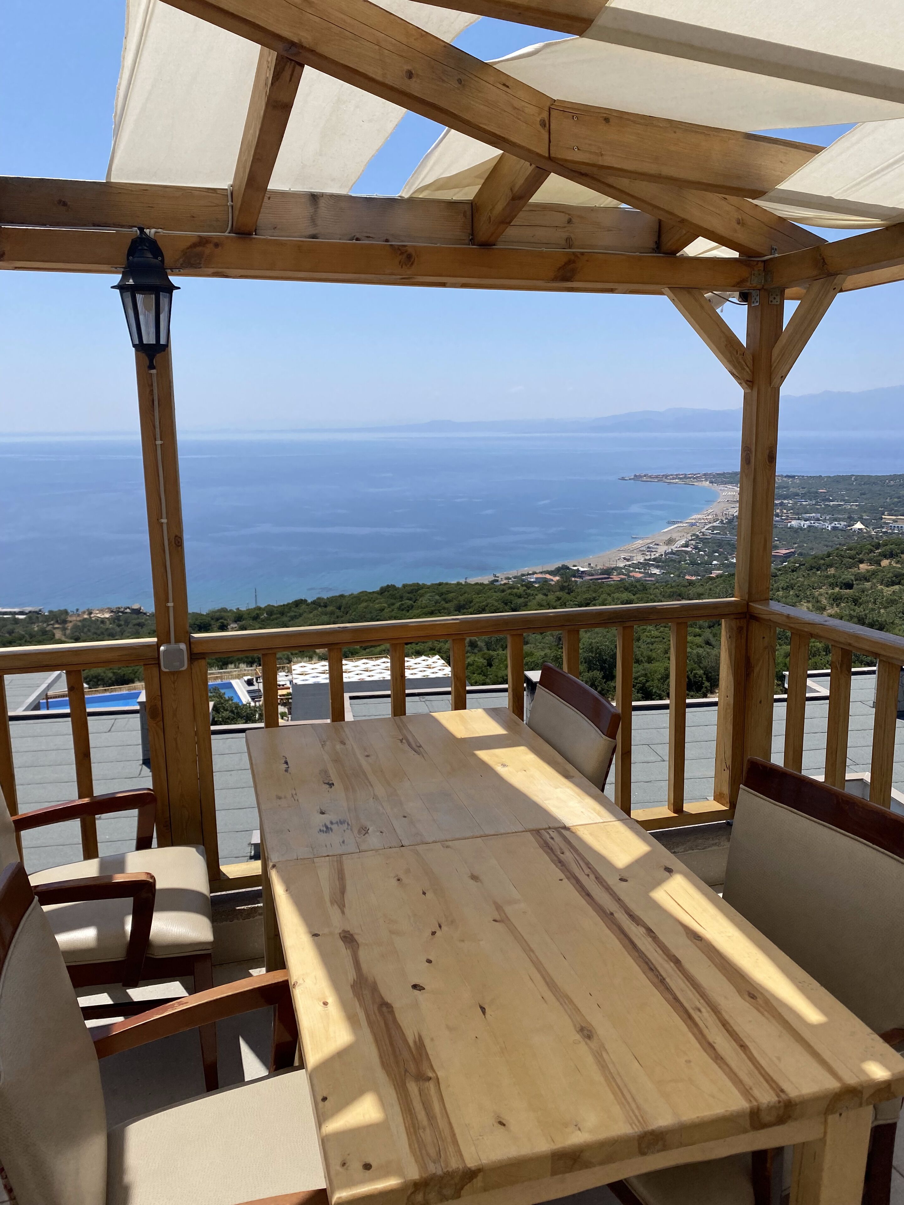 A wooden deck with a table and chairs, overlooking a coastal landscape with buildings and a beach.