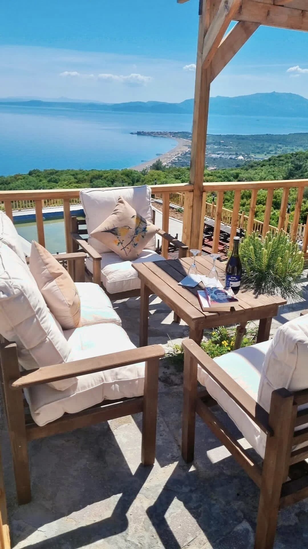 A wooden deck with outdoor seating, a table with a bottle of wine, and a scenic view of a body of water and distant land.