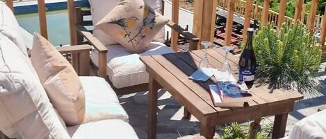 A wooden deck with outdoor seating, a table with a bottle of wine, and a scenic view of a body of water and distant land.