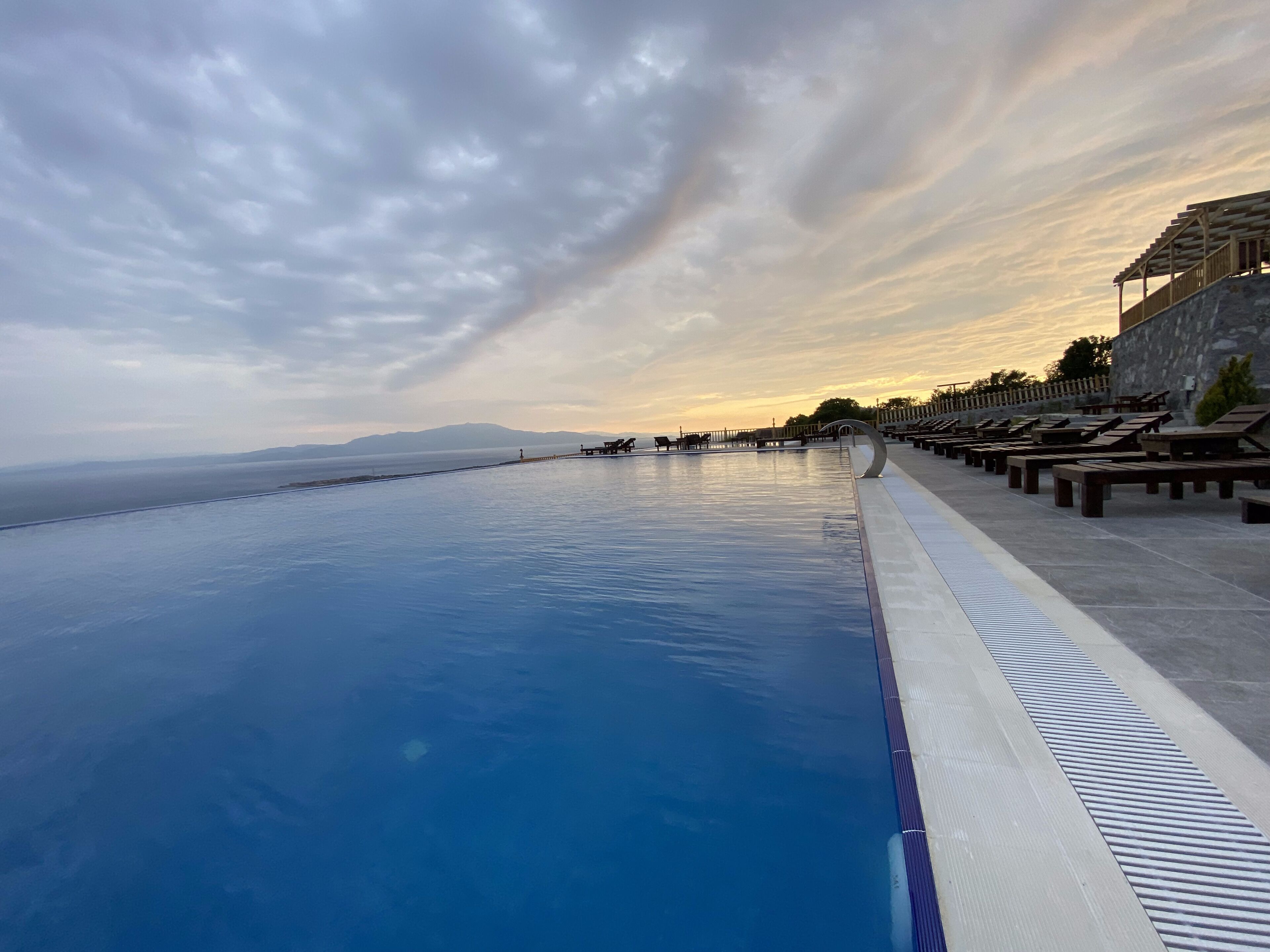 An infinity pool with a view of the sea and mountains, wooden lounge chairs, and a stone wall.