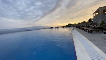 Una piscina infinita con vista al mar y a las montañas, sillones de madera y un muro de piedra.