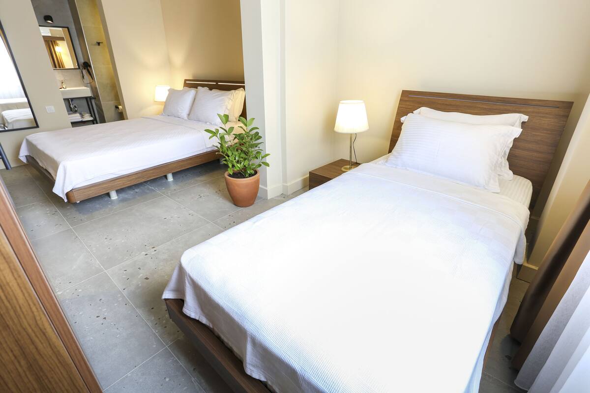 Habitación de hotel con dos camas individuales, una planta en maceta, una mesita de noche con lámpara y un baño con espejo y lavamanos.