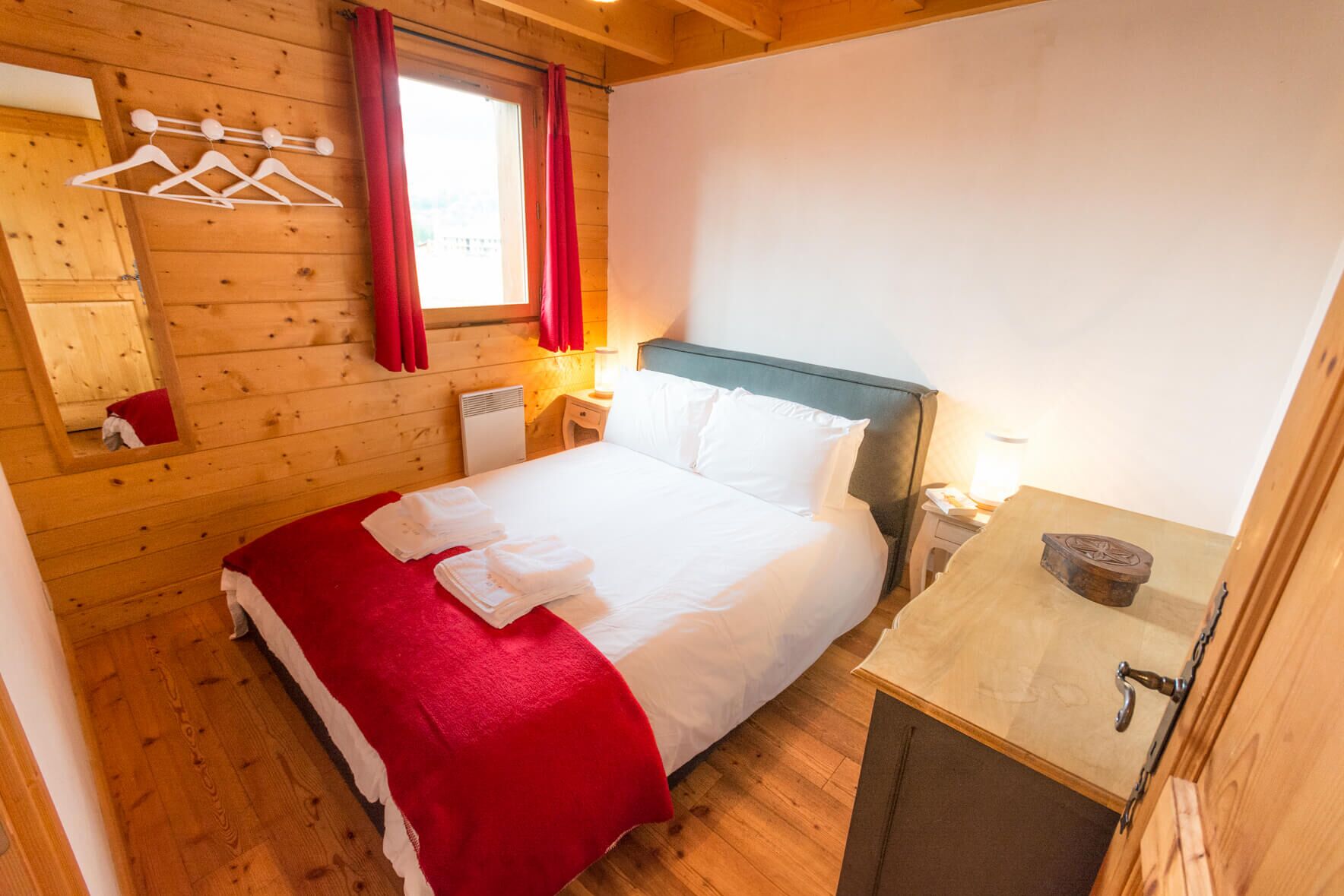 Une pièce en bois avec un lit, une table de chevet, un miroir et une fenêtre ornée de rideaux rouges.