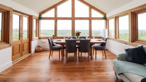 A dining area with a wooden table and chairs, large windows with a view of the countryside, and a wooden floor. - Trevilges Cottage (Helston)