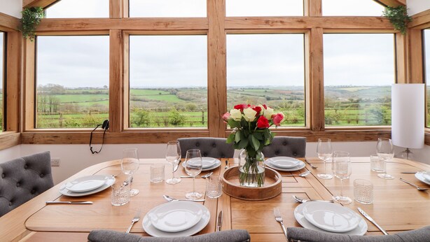 A dining table set for six with a view of a rural landscape through large windows. - Trevilges Cottage (Helston)