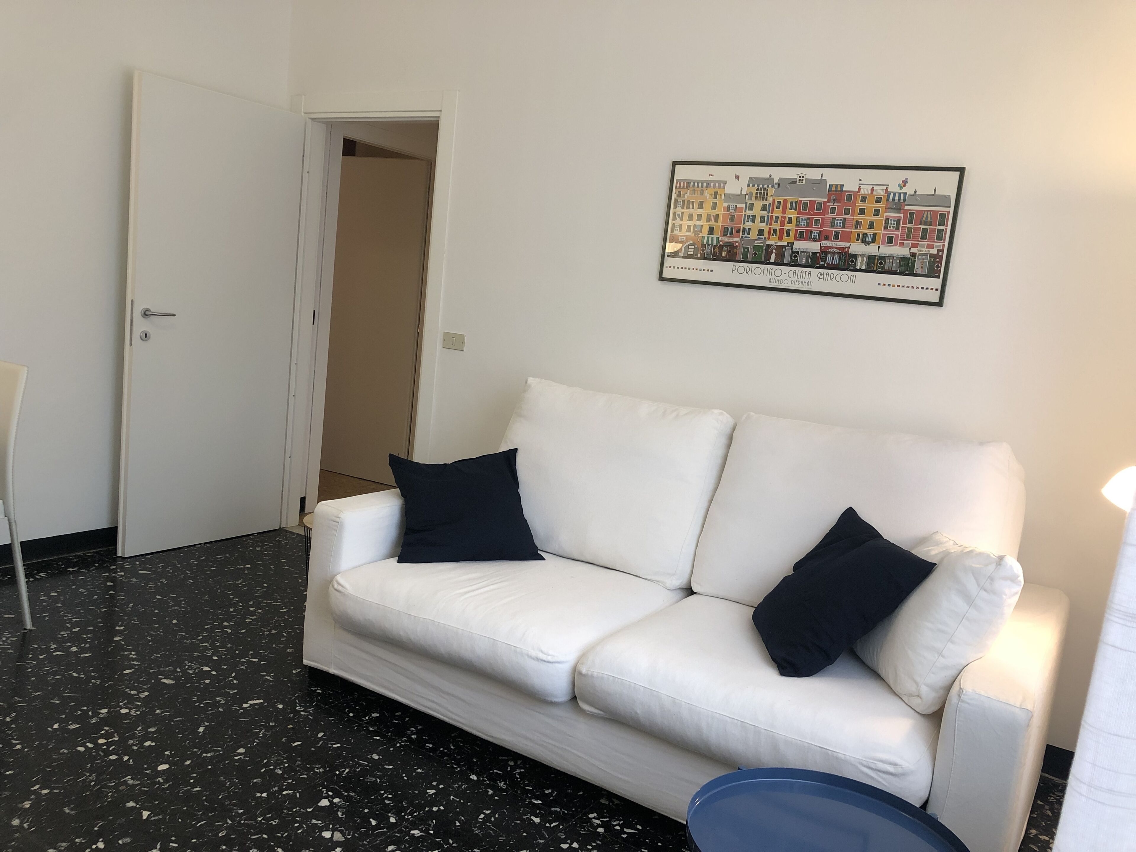 A white sofa with two black cushions, a framed cityscape picture, and a door leading to another room.