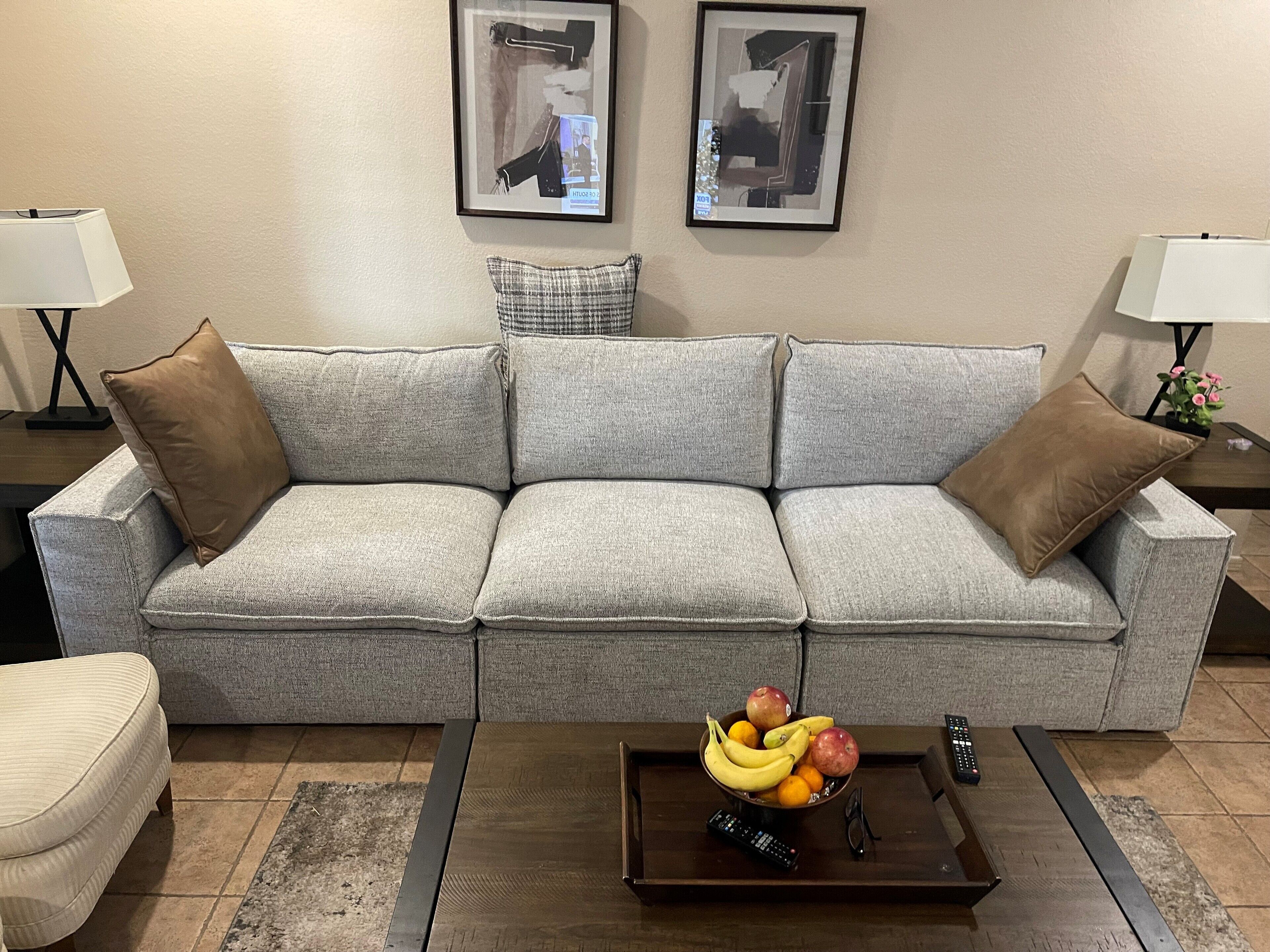 A grey sofa with brown cushions, a wooden coffee table with fruit and a remote control, and framed pictures on the wall.