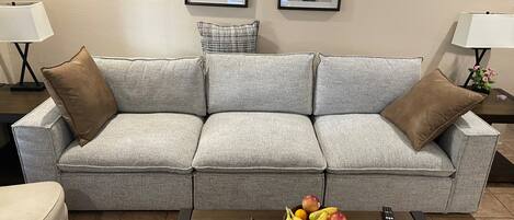 A grey sofa with brown cushions, a wooden coffee table with fruit and a remote control, and framed pictures on the wall.