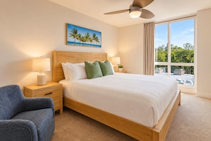 A hotel room with a large bed, a ceiling fan, a painting of palm trees, and a view of greenery through the window. - Whitewater Ocean View Condo | North Coast Village (Oceanside)