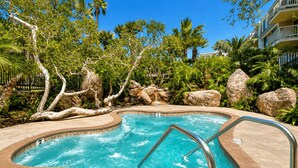 Une piscine à bord incurvé, entourée de rochers et d’arbres, avec un ciel bleu clair au-dessus.