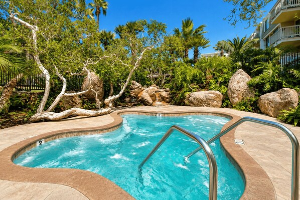 A swimming pool with a curved edge, surrounded by rocks and trees, and a clear blue sky above. - Whitewater Ocean View Condo | North Coast Village (Oceanside)