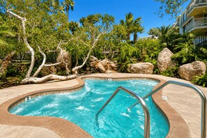 A swimming pool with a curved edge, surrounded by rocks and trees, and a clear blue sky above. - Ocean-View Condo at North Coast Village | B222 (Oceanside)